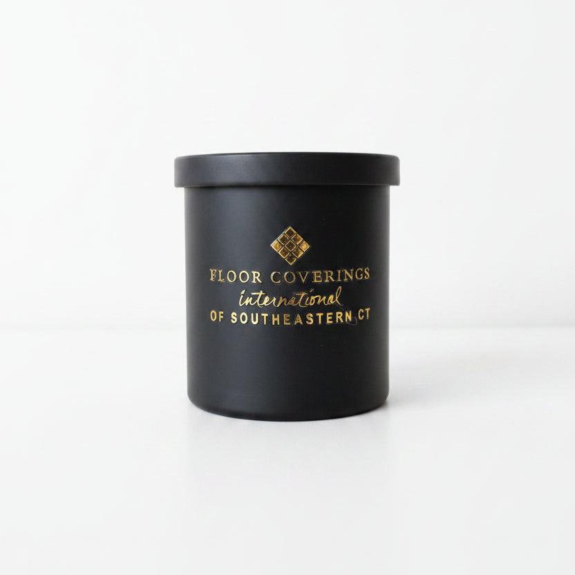 A black cylindrical container with a lid, featuring gold foil printed text that reads 'FLOOR COVERINGS International OF SOUTHEASTERN CT' and a small geometric logo, photographed indoors on a light beige surface.