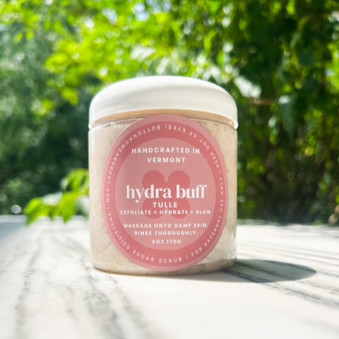 Jar of 'hydra buff' scrub with a pink label on a wooden surface with greenery in the background