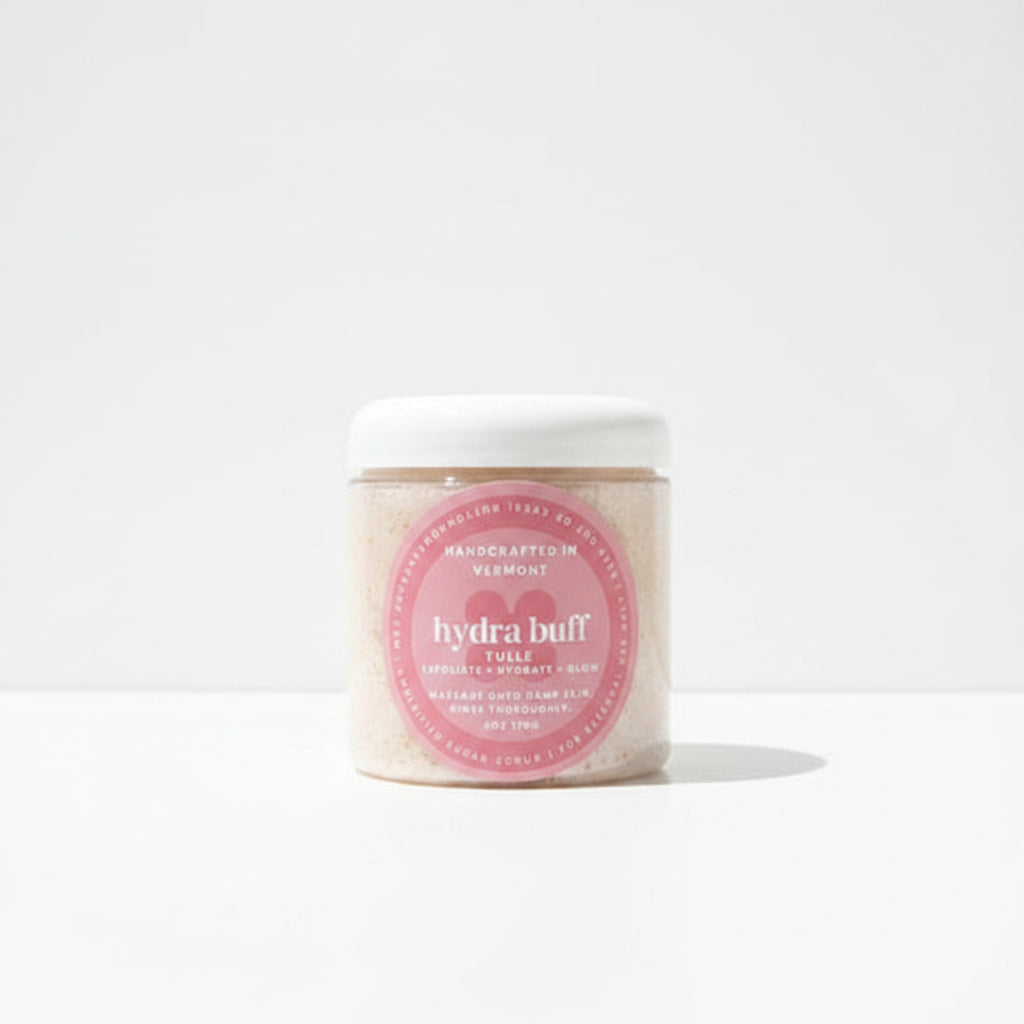 Jar of 'hydra buff' scrub with a pink label on a wooden surface with greenery in the background