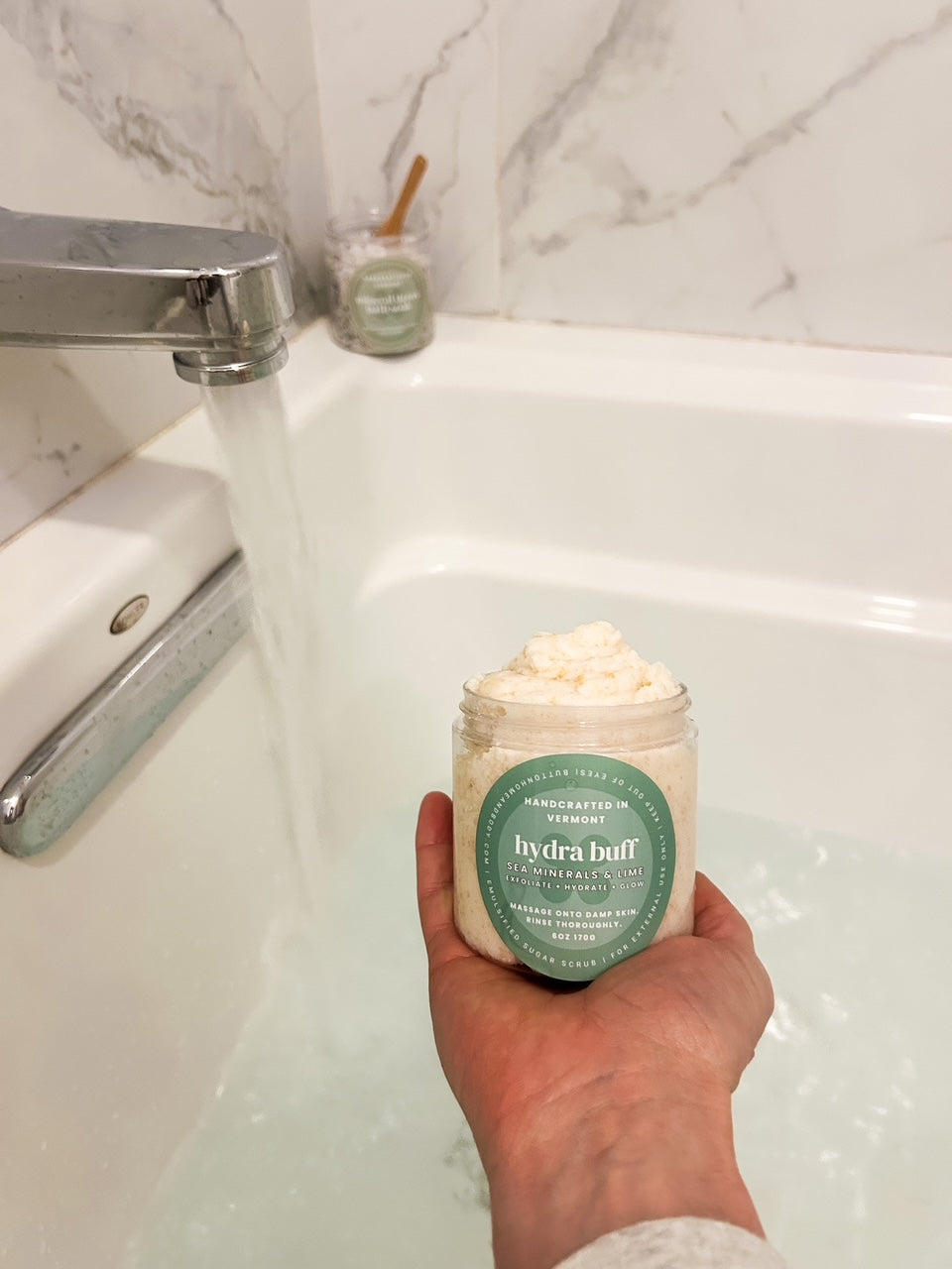 hand holding jar of sugar scrub with water running in a bath tub l