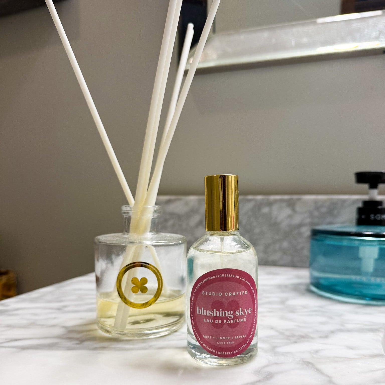 Blushing Skye Eau de Parfum by Button Home & Body in a clear glass bottle with gold cap displayed beside a reed diffuser on a marble countertop.