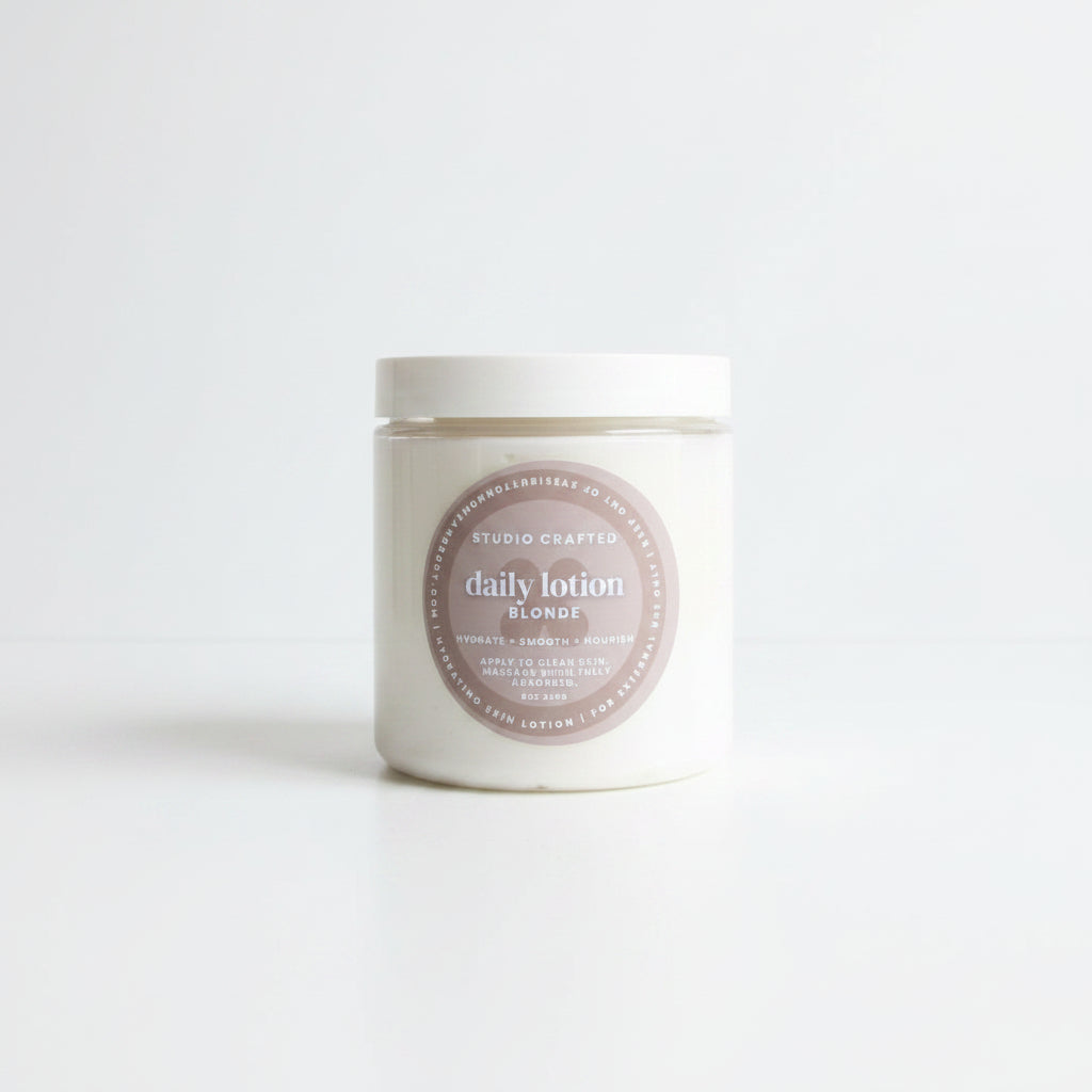 A white jar of Studio Crafted Daily Lotion Blonde sits on a white marble table outdoors, with a patio chair and house siding blurred in the background.