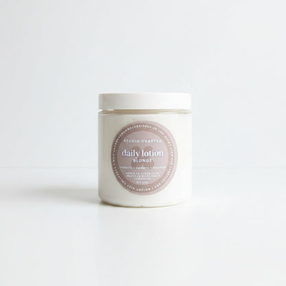 A white jar of Studio Crafted Daily Lotion Blonde sits on a white marble table outdoors, with a patio chair and house siding blurred in the background.