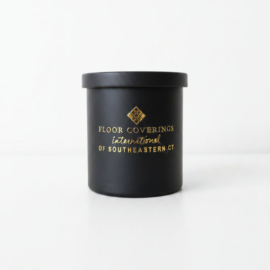 A black cylindrical container with a lid, featuring gold foil printed text that reads 'FLOOR COVERINGS International OF SOUTHEASTERN CT' and a small geometric logo, photographed indoors on a light beige surface.