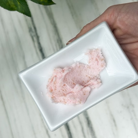 Hand holding a white rectangular dish with slightly shimmering pink sugar scrub highlighting soft consistency on a light marble surface