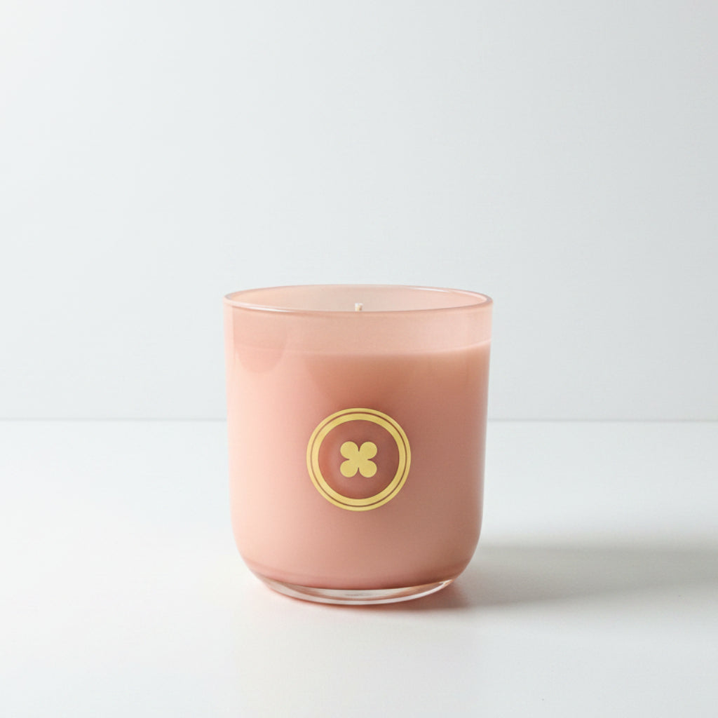 A pink glass candle jar with a gold circular logo on the front, sitting on a wooden windowsill with a window view in the background.