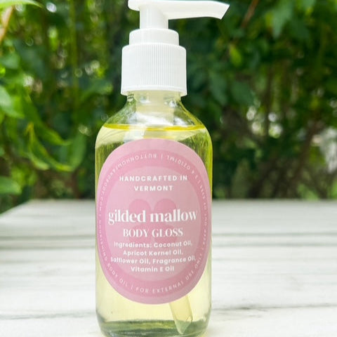 Bottle of gilded mallow body gloss with a pink label on a wooden surface with greenery in the background